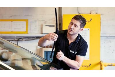 A young man in a black polo shirt with the logo "TRICO" is replacing a windshield wiper blade on a car. He is focused on his task, holding the wiper blade with both hands while ensuring it is properly attached to the car's windshield. 